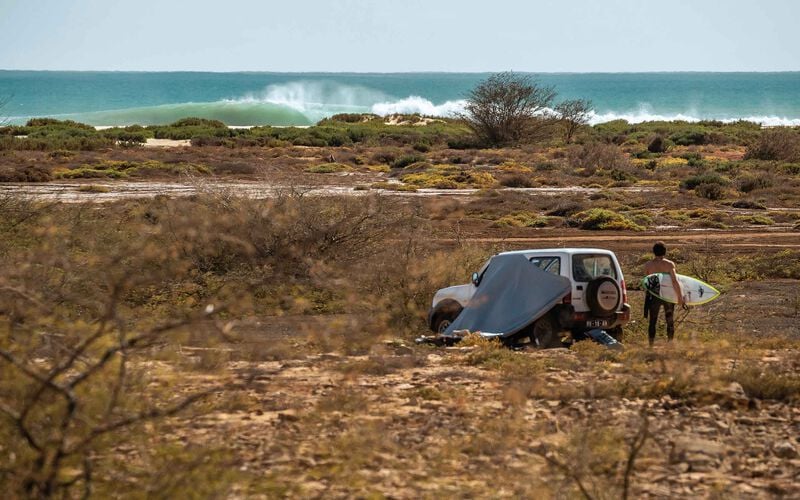 A car, tent and a person with a surfboard in front of a breaking wave. 
