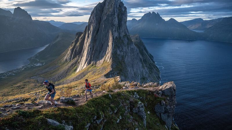 Two runners climb a trail near a tower of rock surrounded by water. 